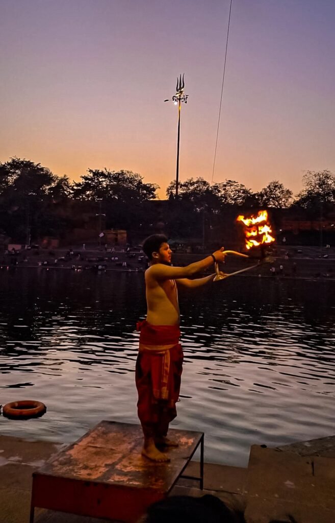 A priest raising a flaming lamp during the evening Shipra aarti at Ujjain, with the river and trees behind.