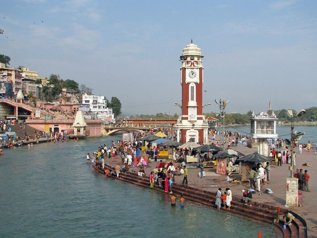 The red clock tower beside Brahmakund at Har Ki Pauri, with pilgrims on the ghat steps and the Ganga flowing past.