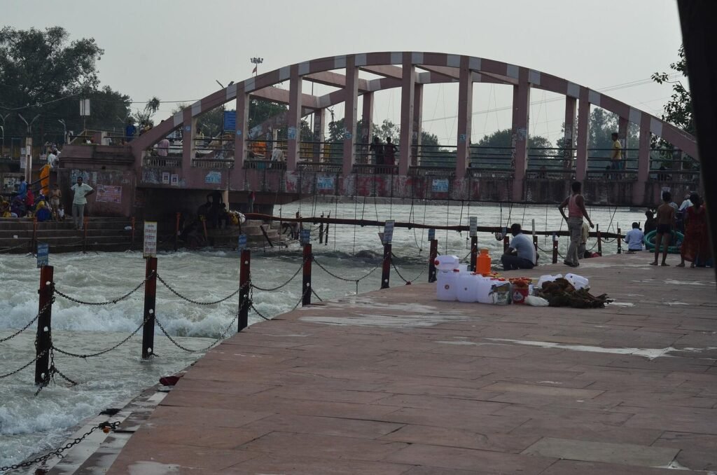 An arched footbridge over the Ganga at Har Ki Pauri, with a chained ghat edge in the foreground.