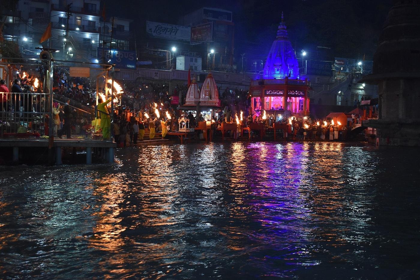 Rows of aarti lamps glowing at Har Ki Pauri after dark, reflected in the Ganga with a lit temple behind.