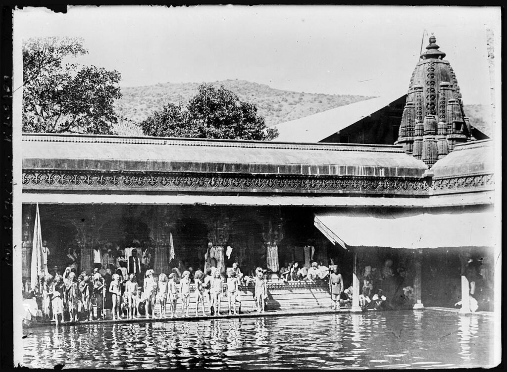 A 1928 black and white photograph of Kushavarta Kund at Trimbak, with people at the water's edge below the temple spire and roofs.