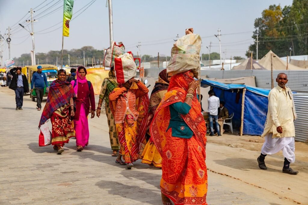Women pilgrims in bright saris carrying bundles on their heads along a camp road at the Kumbh Mela, Prayagraj.