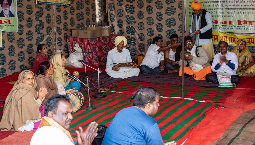 Devotees seated on rugs under a printed cloth pandal at a Kumbh Mela camp in Prayagraj, singing with cymbals and a drum at a satsang.
