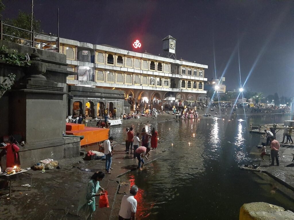 Ramkund on the Godavari after dark, lit by floodlights, with pilgrims at the water's edge and the old arcade behind.