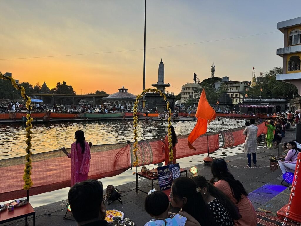 People gathered on the ghat at Ramkund at sunset, with flags and the Godavari glowing orange.