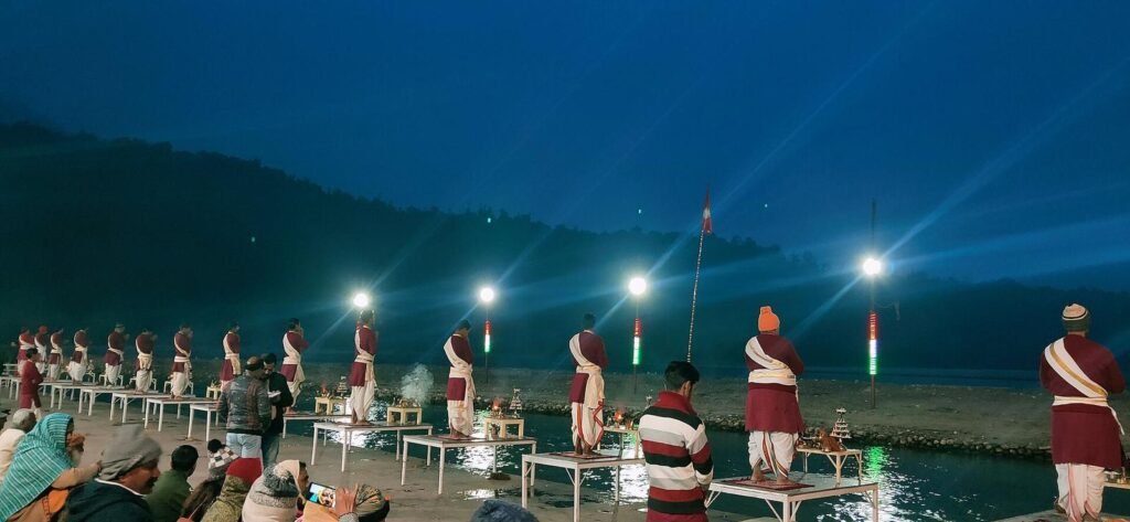 Priests in matching dress performing the evening Ganga aarti at Triveni Ghat, Rishikesh, with lights along the river.
