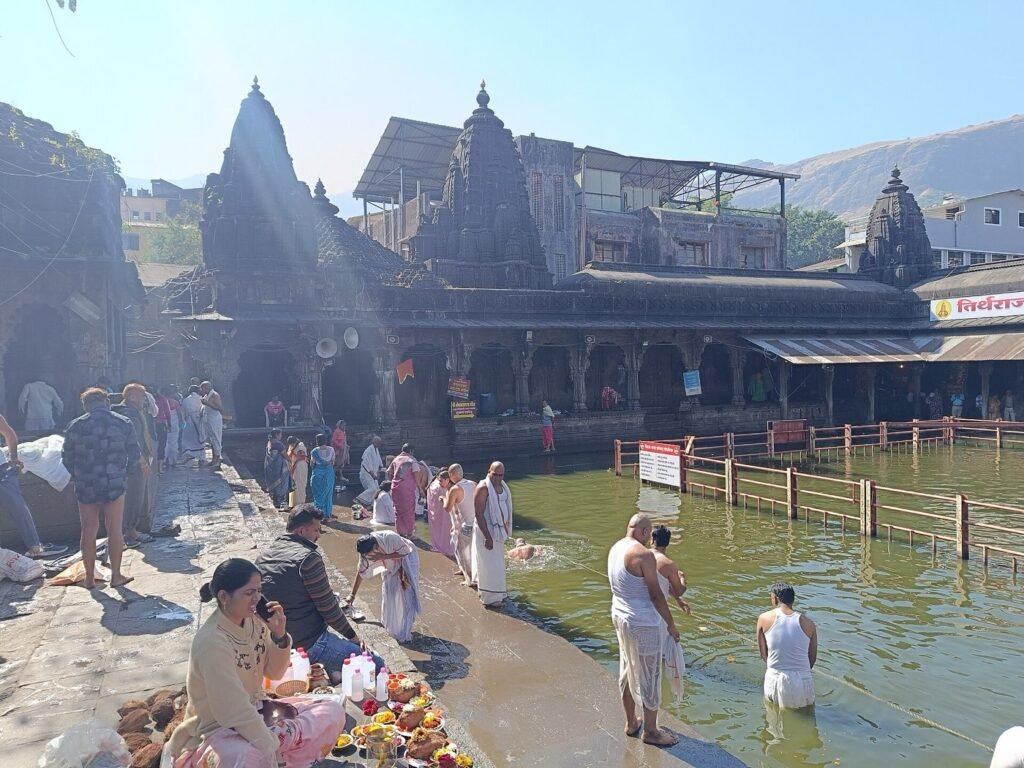 Pilgrims at the edge of the Kushavarta kund in Trimbakeshwar on a sunny day, with the stone colonnade and temple spires around the tank.