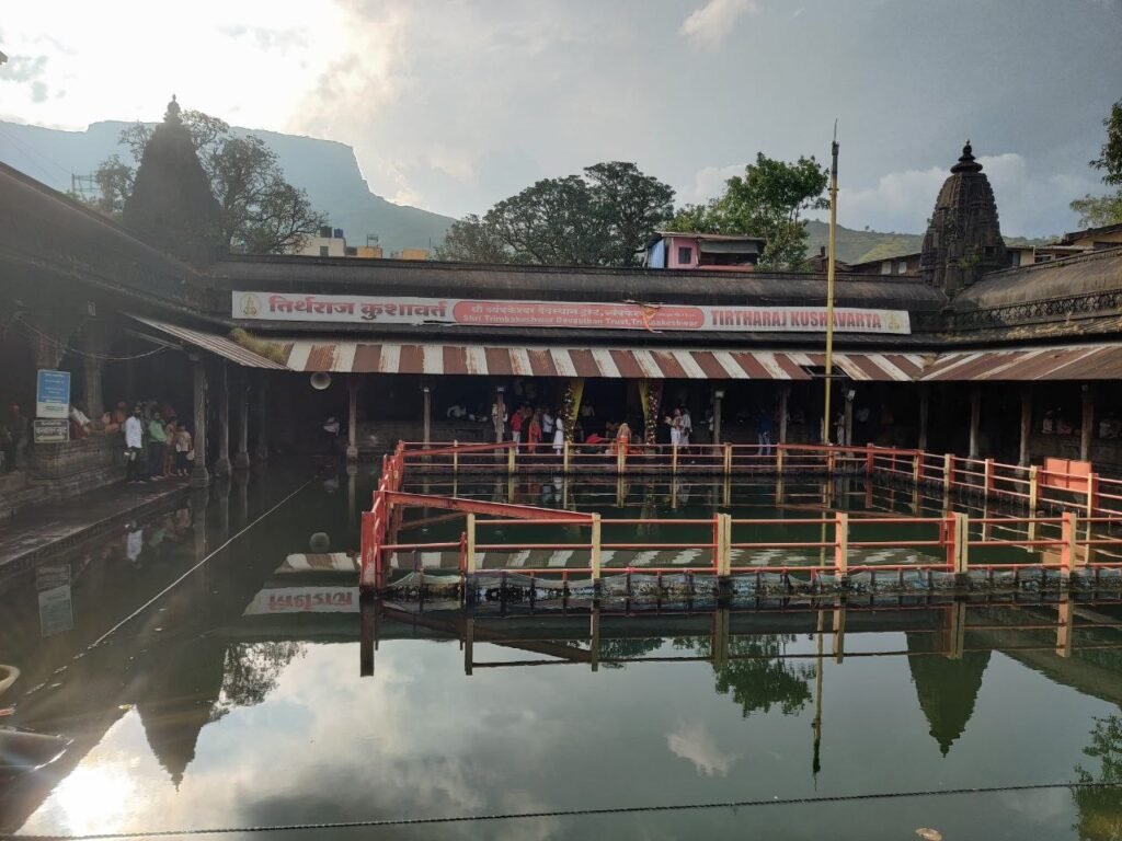 The Kushavarta kund in Trimbakeshwar: a stone tank with railings in still water, the colonnade and temple spires around it and hills behind, reflected in the water.