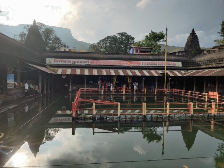 The Kushavarta kund in Trimbakeshwar: a stone tank with railings in still water, the colonnade and temple spires around it and hills behind, reflected in the water.
