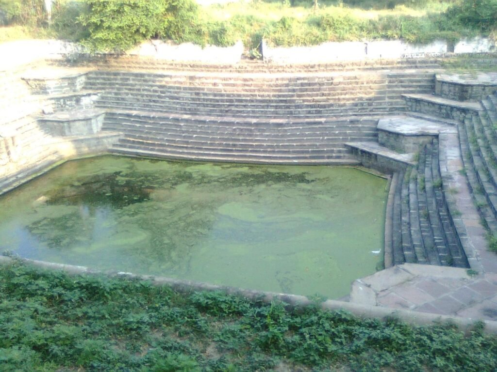 The stone-stepped water tank (kund) at Maharshi Sandipani Ashram, Ujjain, with green water and grass around it.